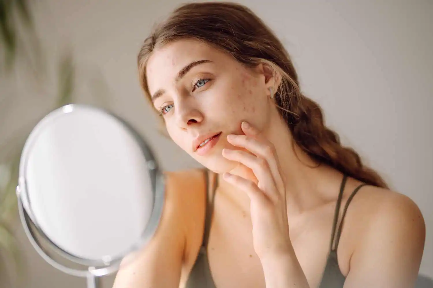 Young woman inspecting acne and blemishes on her cheek while looking in a mirror